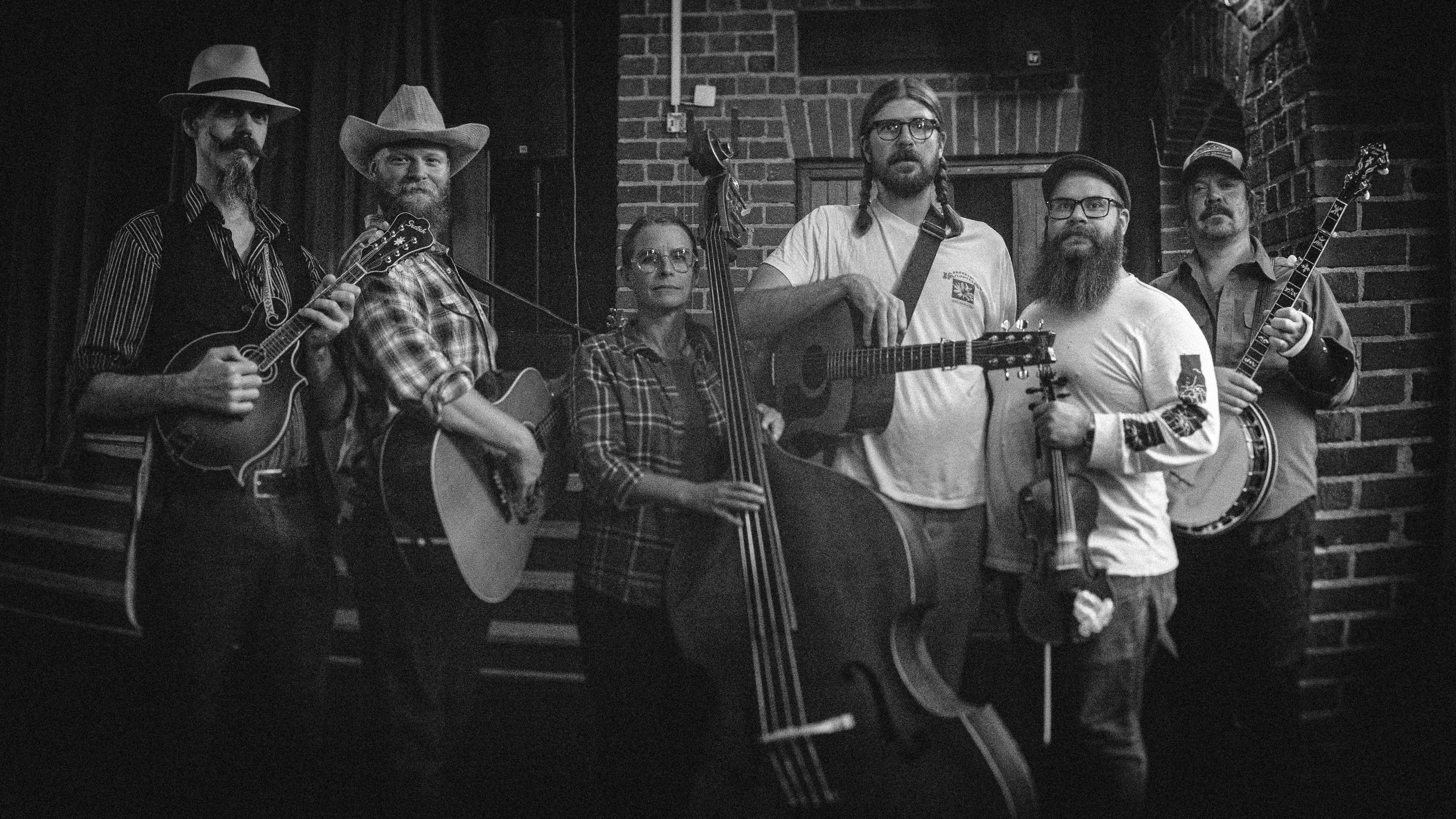 Five men holding mandolin, guitars, fiddle and banjo standing around one woman holding a double bass